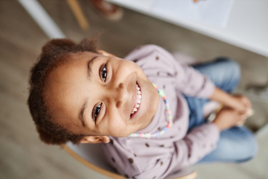 Above shot of face of happy little African American learner of nursery school sitting by table and looking at camera with smile